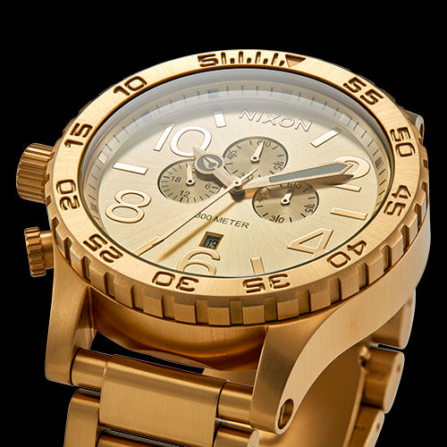 A close-up shot of a luxurious gold Nixon chronograph watch featuring a textured dial, three sub-dials, a date window, and a matching gold link bracelet, set against a dark background.